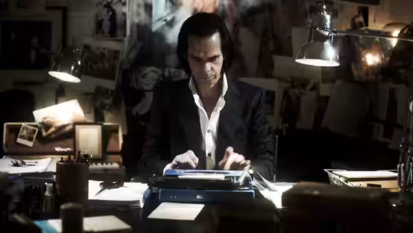 Nick Cave typing at a cluttered desk surrounded by photographs