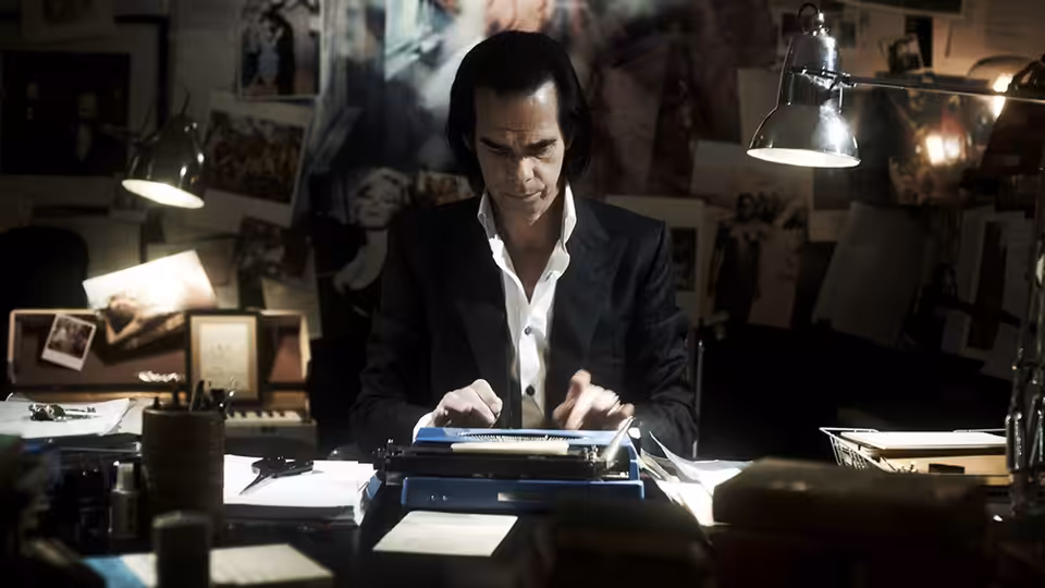 Nick Cave typing at a cluttered desk surrounded by photographs