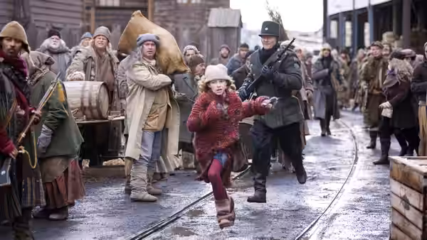 Lyra Belacqua runs through a crowded street alongside an armed guard
