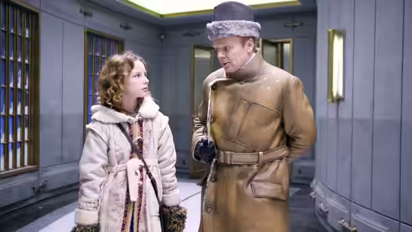 Lyra talks with a uniformed man in a fur hat inside a corridor