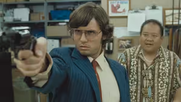 Mark Chapman (Jonas Ball) points a handgun inside a cluttered shop as a man watches