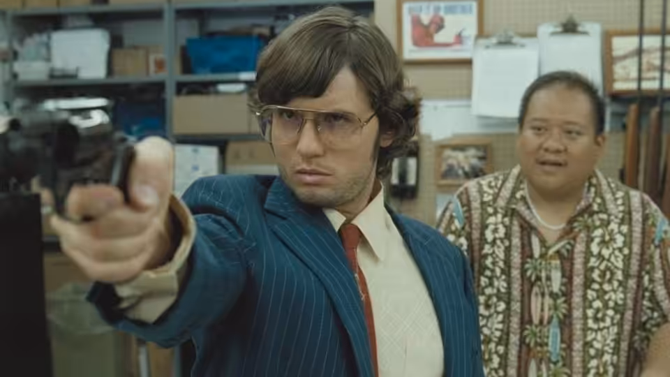 Mark Chapman (Jonas Ball) points a handgun inside a cluttered shop as a man watches