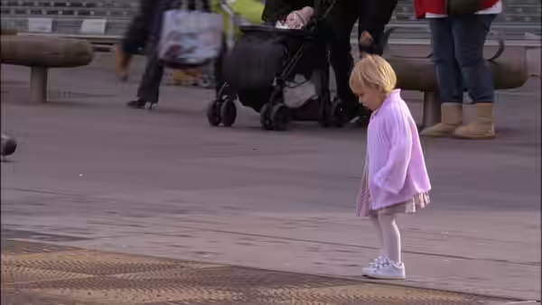 A little girl in a pink shawl stands on a paved plaza near strollers