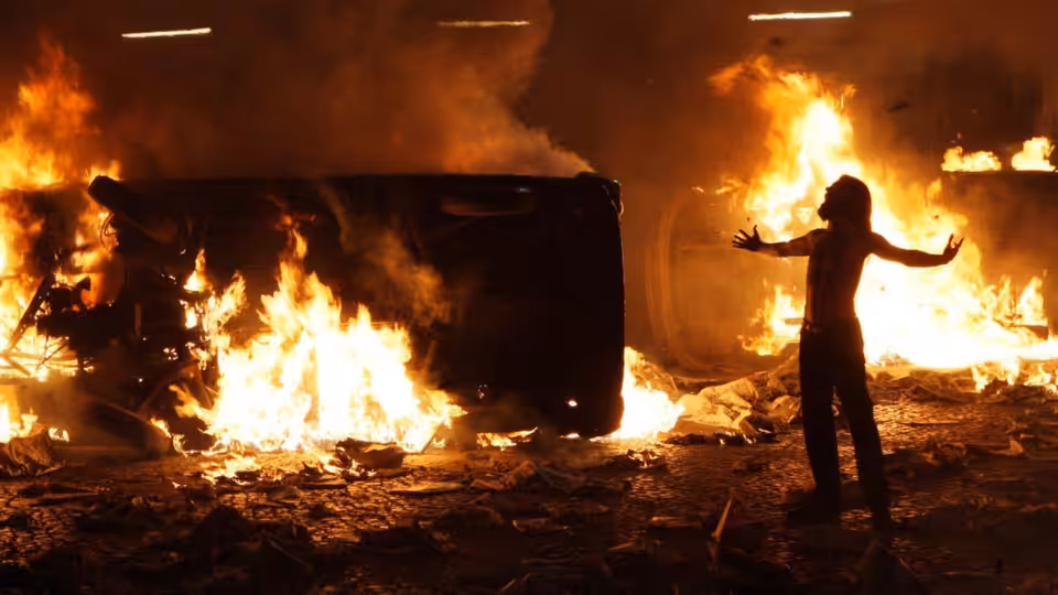 A silhouetted figure spreads their arms beside a van engulfed in flames