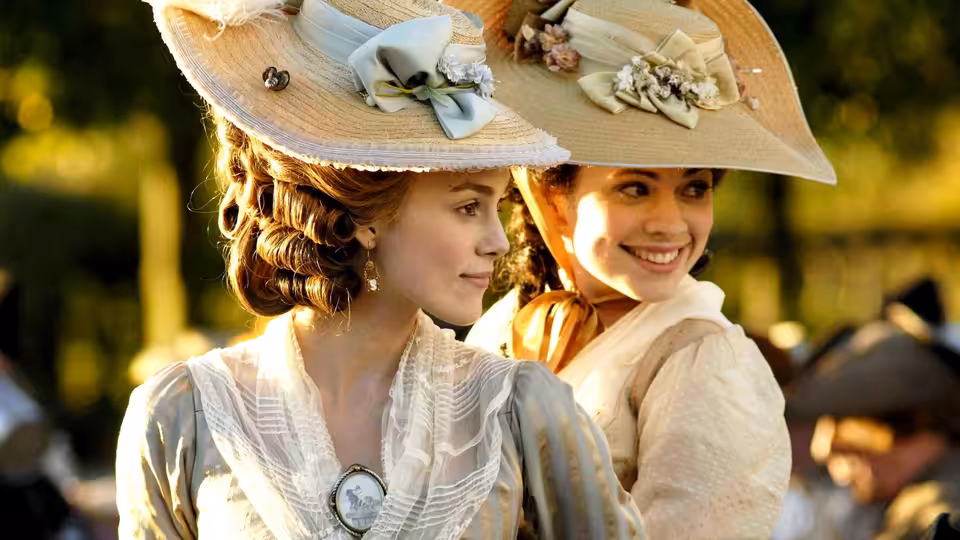 Georgiana Cavendish (Keira Knightley) and Bess Foster (Hayley Atwell) smile together outdoors in elaborate hats