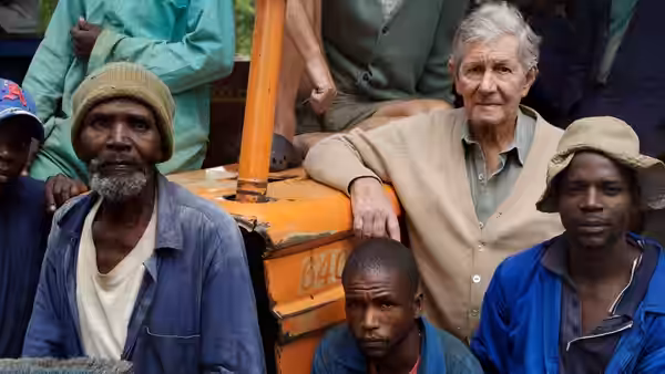 An older white man sits beside Black farm workers around an orange tractor