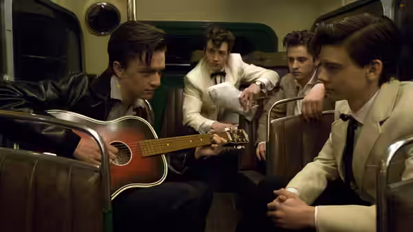 John Lennon (Aaron Taylor-Johnson) plays guitar while three young men sit around him on a train