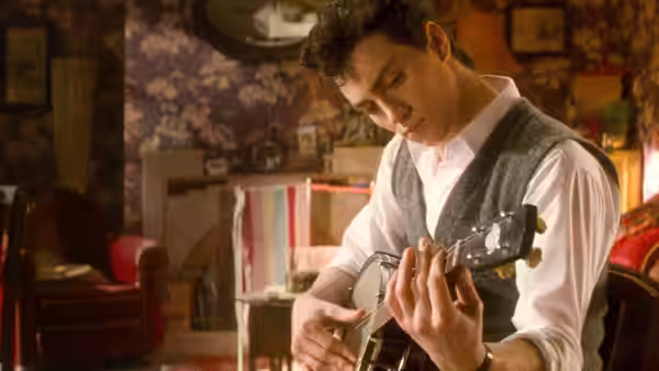 A young man in a white shirt and vest plays guitar in a living room