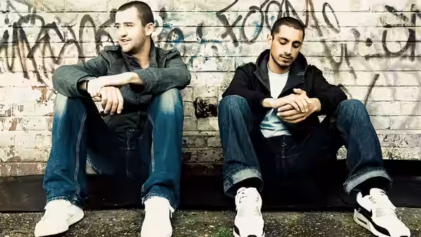 Chris (Daniel Mays) and Shifty (Riz Ahmed) sit against a graffiti-covered wall