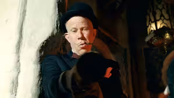 The Devil (Tom Waits), wearing a bowler hat, points toward someone offscreen