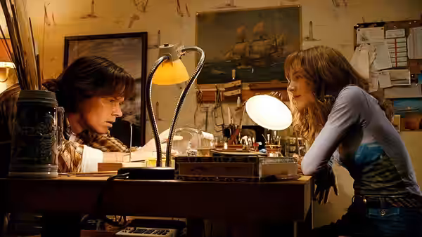 A young woman leans across a cluttered desk toward a dark-haired man
