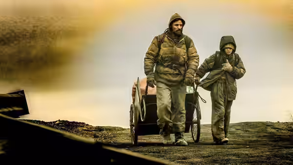 The Father and Boy walk beside their handcart across a barren landscape