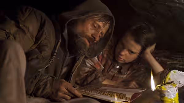 The Father and Boy read together by candlelight inside a dark shelter