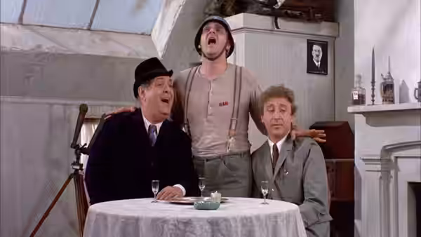 Max Bialystock sits at a table as a man sings behind him and another watches