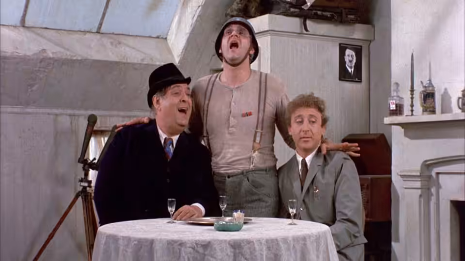 Max Bialystock sits at a table as a man sings behind him and another watches