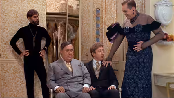 Max Bialystock and Leo Bloom sit as a man in a dress stands beside them