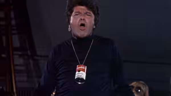 A man in a black turtleneck and identification badge sings on a dark stage