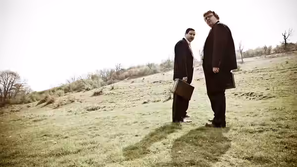 Two men in dark coats stand on a grassy hillside, holding briefcases