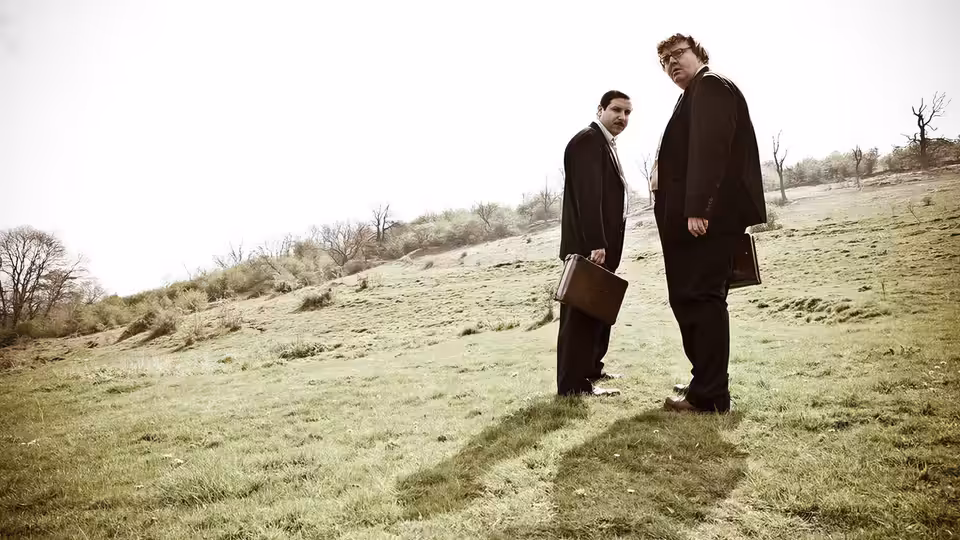Two men in dark coats stand on a grassy hillside, holding briefcases