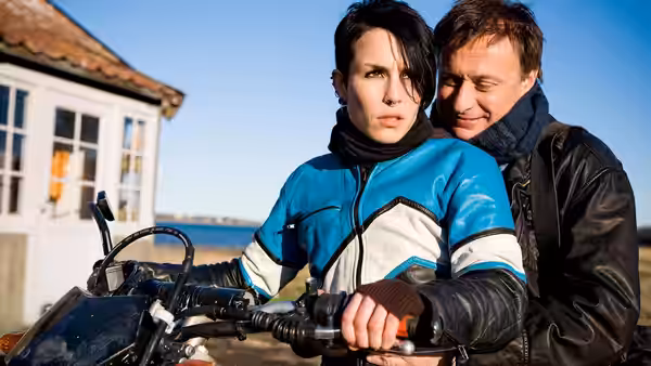 Lisbeth Salander (Noomi Rapace) sits at a motorcycle as Mikael Blomkvist (Michael Nyqvist) leans behind her