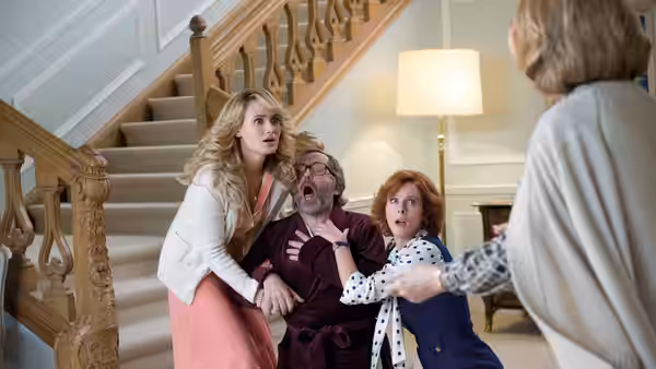 A blonde woman and a red-haired woman steady a startled man in the grand staircase hall