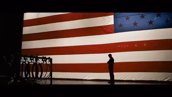 A lone man stands before a giant American flag beside a bank of microphones
