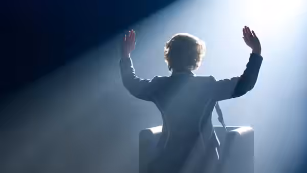 A woman at a lectern raises both hands toward a bright spotlight