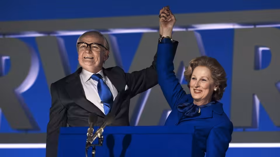 Margaret Thatcher (Meryl Streep) raises a clasped hand beside a suited man at a campaign podium