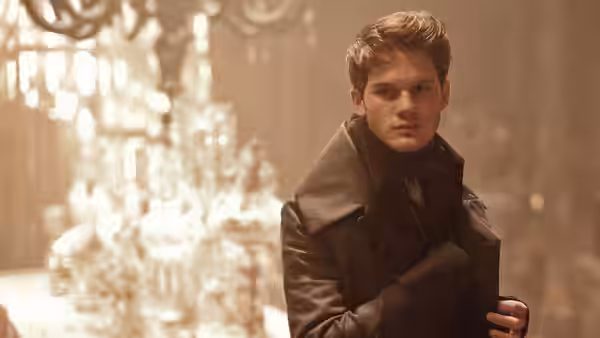 Pip (Jeremy Irvine) stands beneath a chandelier, clutching his coat in an ornate room