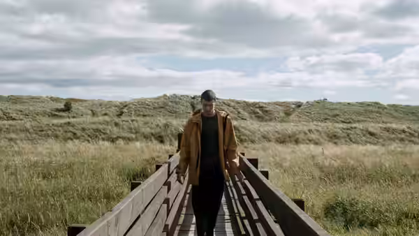 A young man walks along a wooden boardwalk through grassy dunes