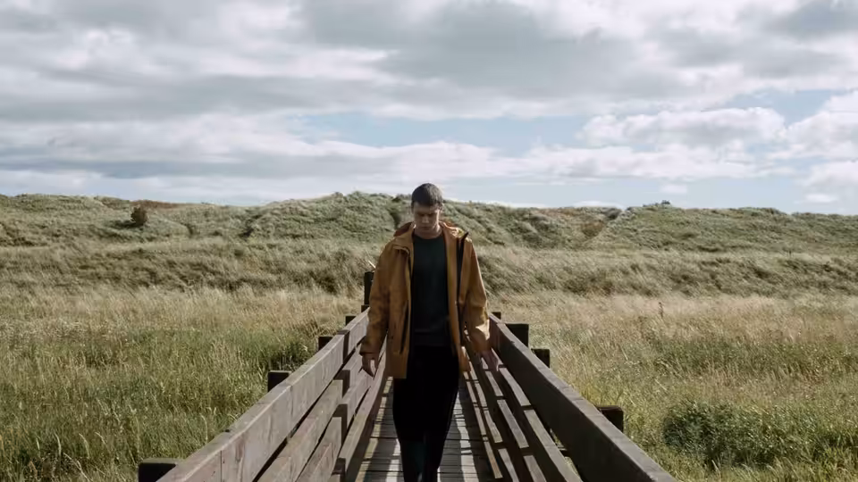A young man walks along a wooden boardwalk through grassy dunes
