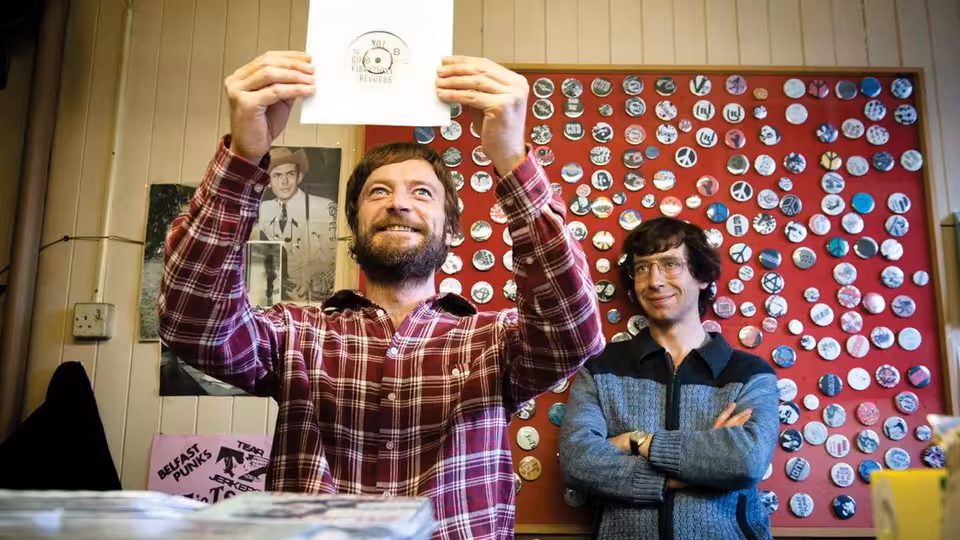 Terri Hooley (Richard Dormer) holds up a record sleeve inside his shop as a man watches