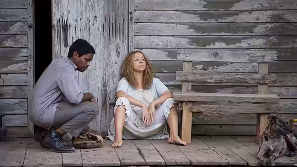 A young man crouches beside a barefoot woman sitting outside a weathered wooden house