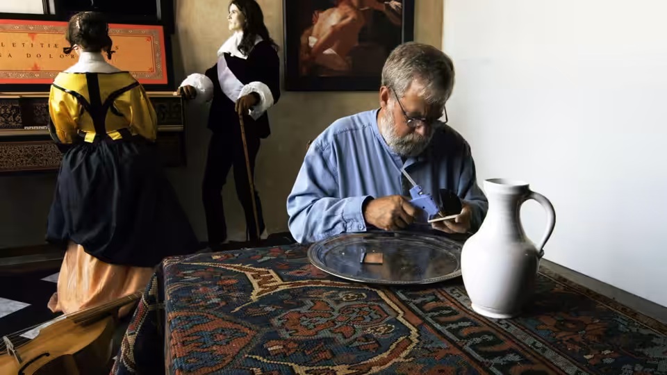 Tim Jenison works at a table beside figures arranged in a painted interior