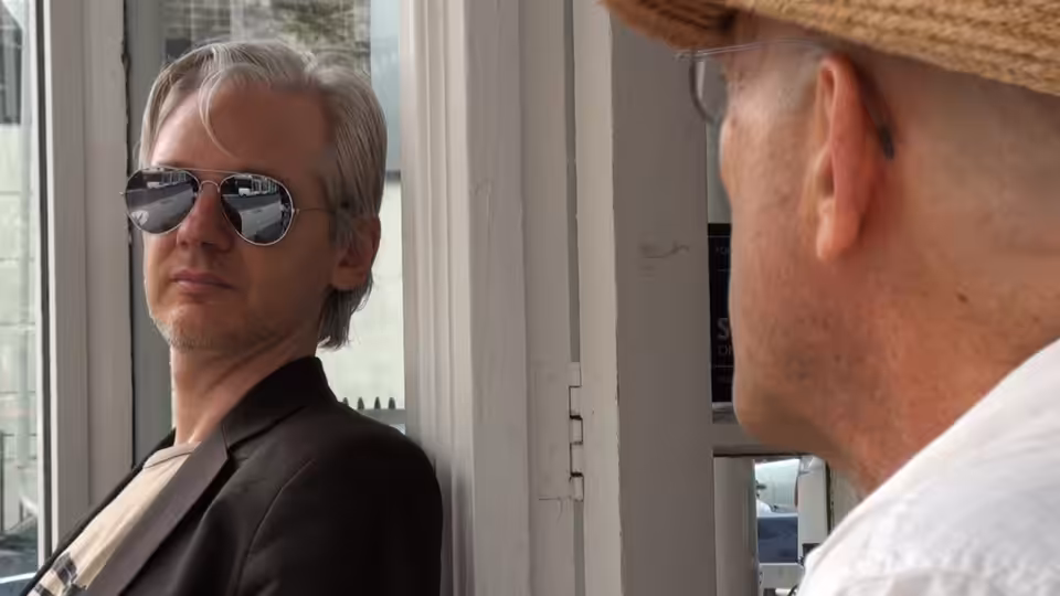 Julian Assange in sunglasses faces a man beside a window