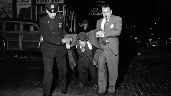 A uniformed officer and a suited man escort a shorter man along a city street