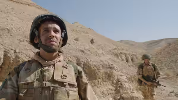 A helmeted soldier stands in a rocky valley as another approaches with a rifle