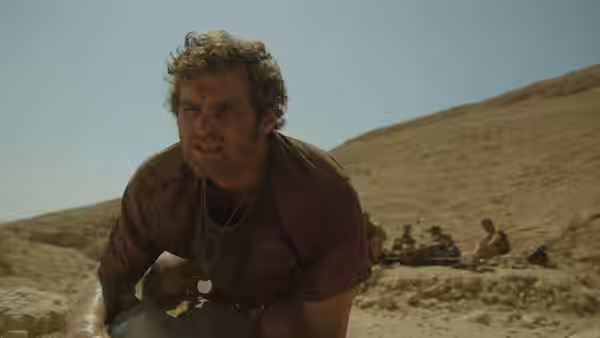 A curly-haired soldier crouches on rocky ground as comrades gather behind him