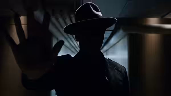 A shadowed man in a wide-brimmed hat reaches toward the viewer
