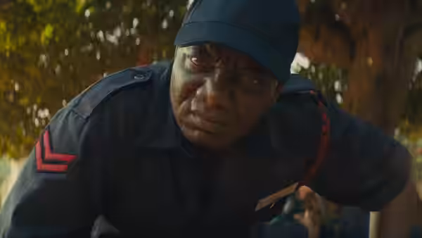 A uniformed man crouches beneath the trees, looking intently to one side