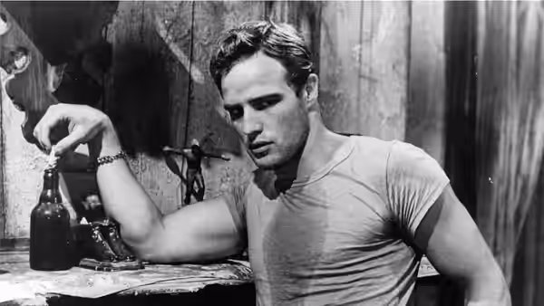 Marlon Brando as Stanley Kowalski leans over a table, holding a bottle in a bare room