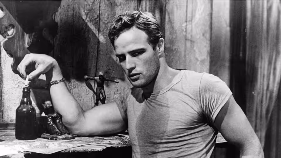 Marlon Brando as Stanley Kowalski leans over a table, holding a bottle in a bare room