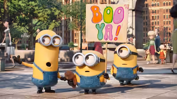 Three Minions walk through a city square, one carrying a colorful protest sign