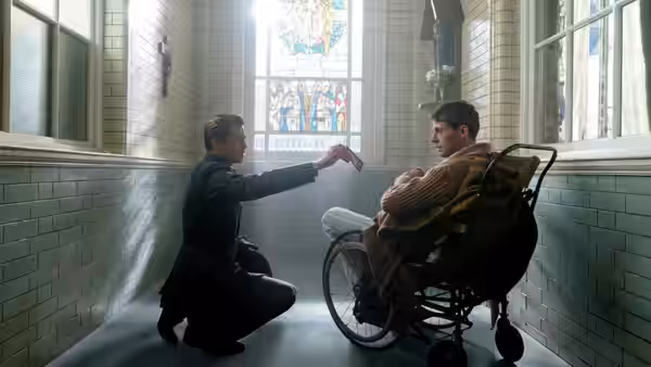 Max Vatan (Brad Pitt) kneels beside a man seated in a wheelchair in a tiled corridor