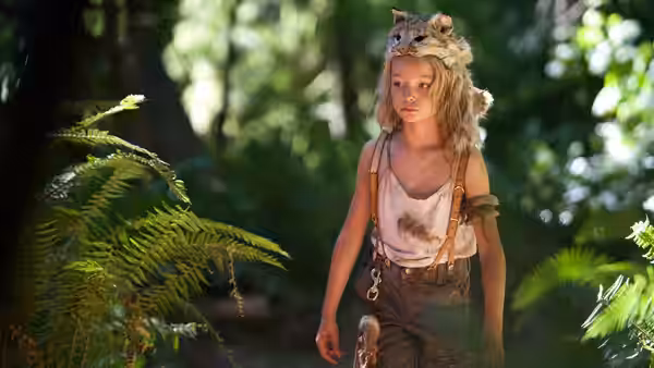 A young girl wearing animal bones and a fur headdress walks through the forest