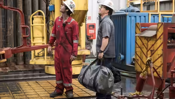 Two oil-rig workers in hard hats stand among pipes and equipment