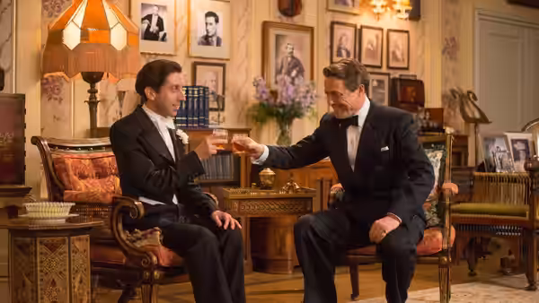 Cosmé McMoon (Simon Helberg) and St. Clair Bayfield (Hugh Grant) toast in a furnished sitting room