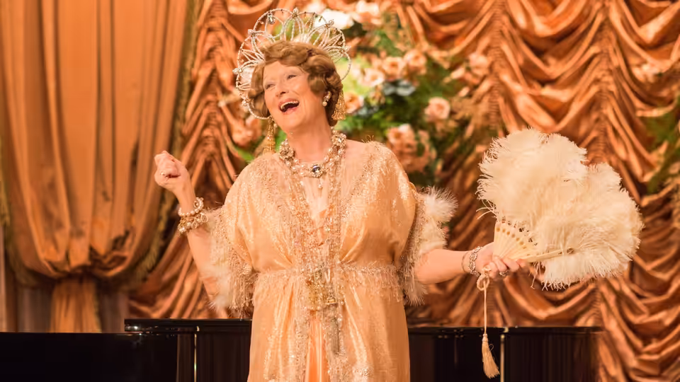 Florence Foster Jenkins (Meryl Streep) sings onstage, holding a feather fan before ornate curtains