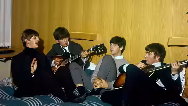 The Beatles sit together in a room, two holding guitars as another gestures