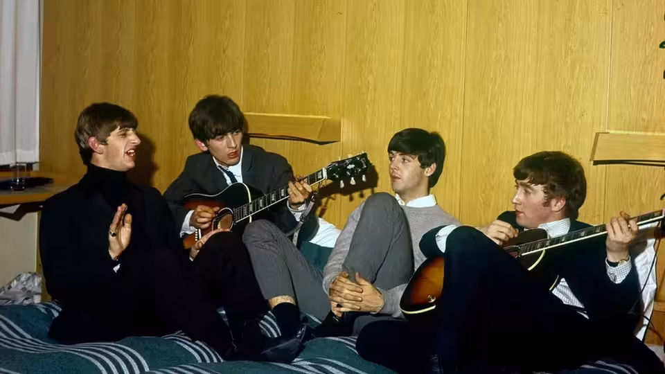 The Beatles sit together in a room, two holding guitars as another gestures